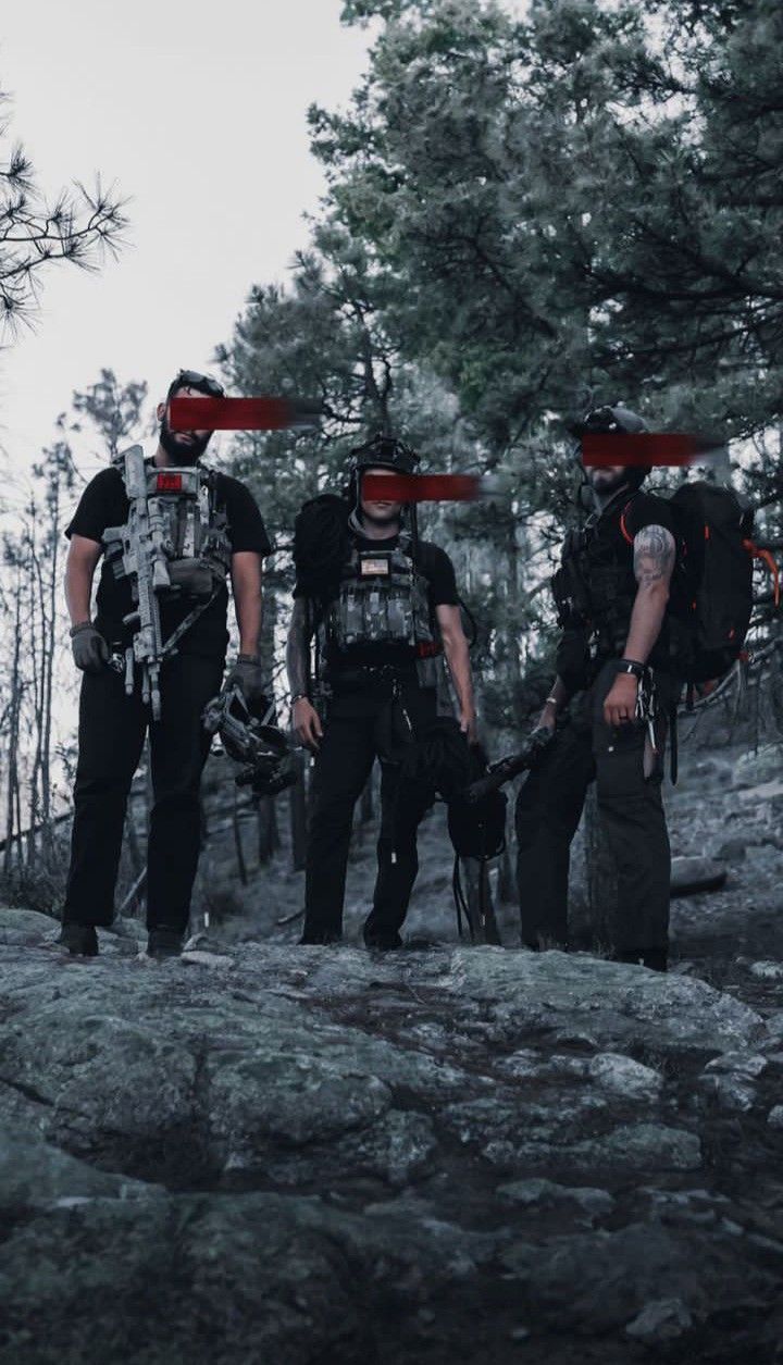 Three clothed men in field gear together in a pine forest.